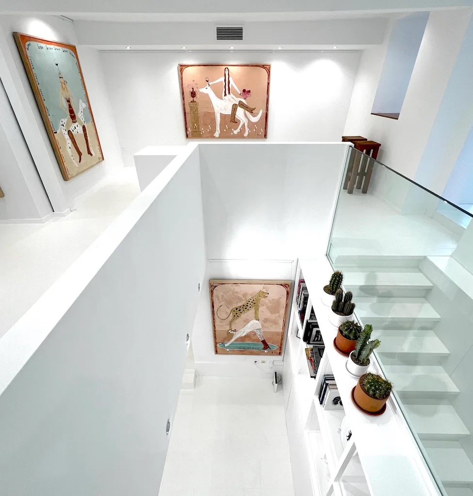 Vue plongeante sur l'accrochage de JAGAL : les œuvres réparties sur deux niveaux, l'escalier de verre, les plantes.